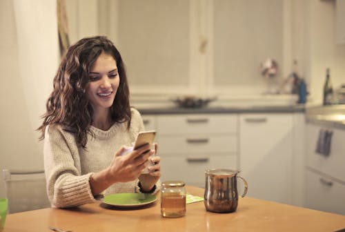 Cheerful young female in casual wear chatting with friends on smartphone while sitting at table and drinking tea in modern kitchen