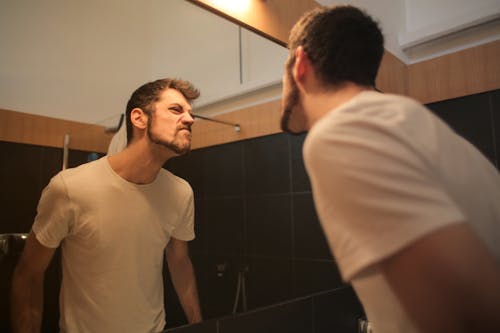 Low angle side view of young bearded male in casual shirt standing in bathroom and looking at with frown mirror in morning