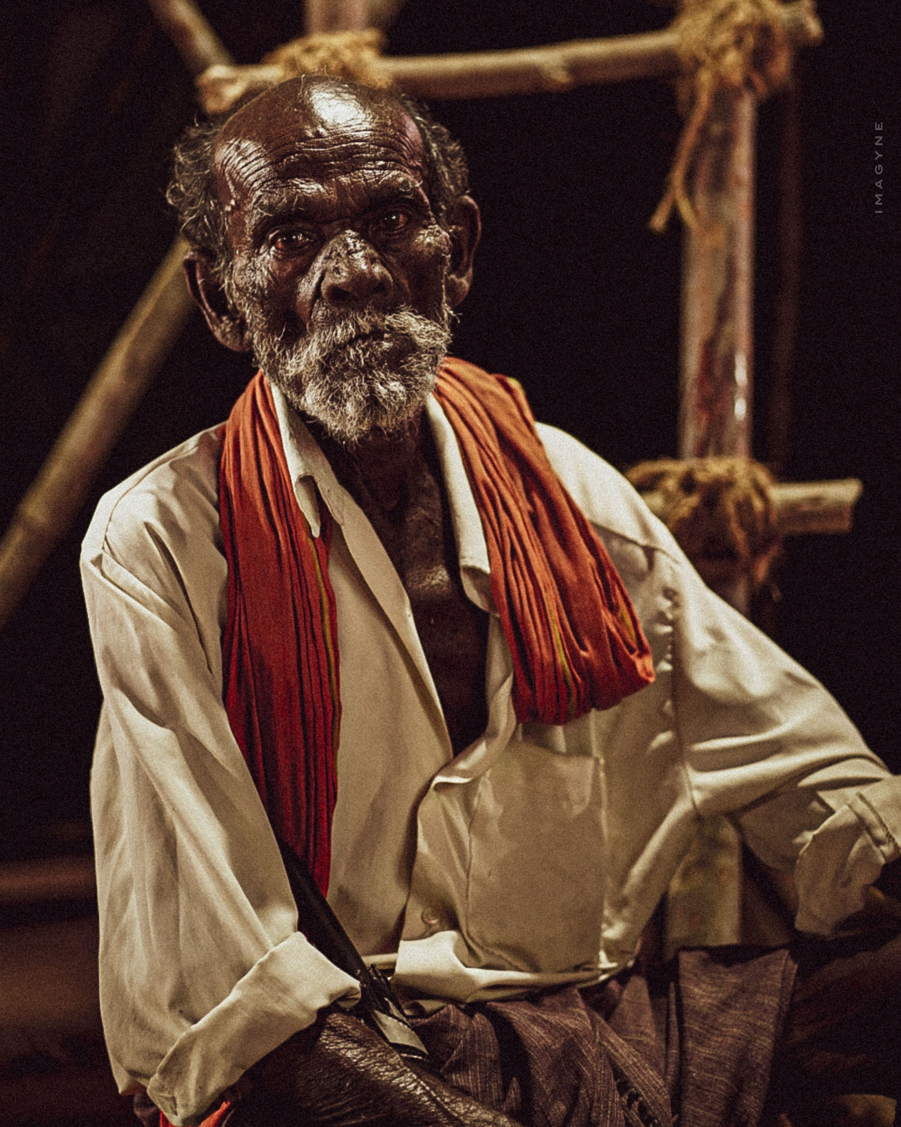 Portrait of an elderly man wearing a red scarf, seated indoors in rustic surroundings.