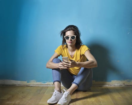 Woman in sunglasses sitting against a blue wall texting on her smartphone.