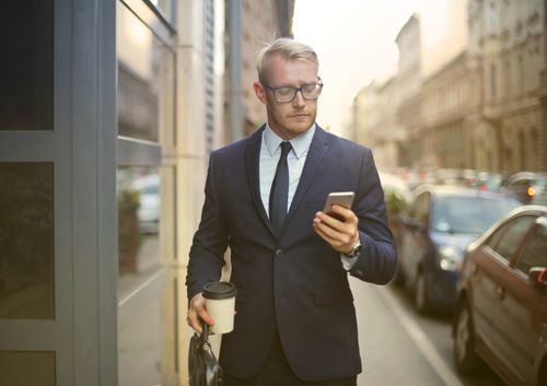 Free Professional businessman in formal wear walking on a city street while texting on his phone and holding coffee. Stock Photo