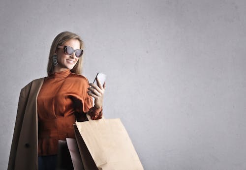 Free Smiling female in trendy wear and stylish sunglasses standing with paper shopping bags on background of gray wall and browsing mobile phone Stock Photo