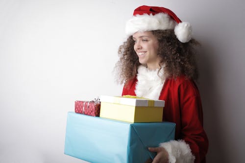 Side view of cheerful female with curly hair wearing Santa hat and costume standing with stack of wrapped presents on white background in studio and looking away