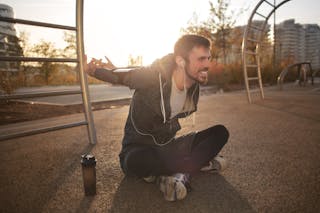 Side view of delighted sportsman in activewear sitting with crossed legs on ground and stretching arms while training on street and listening to music in earphones