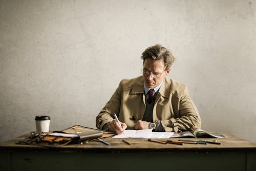 Concentrated male entrepreneur wearing classy jacket and eyeglasses sitting at table with papers and writing supplies while working on blueprint of project