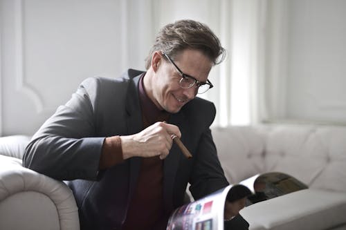 Free Smiling rich gentleman wearing classy suit and stylish eyeglasses sitting with cigar on leather sofa in luxury living room and reading magazine Stock Photo