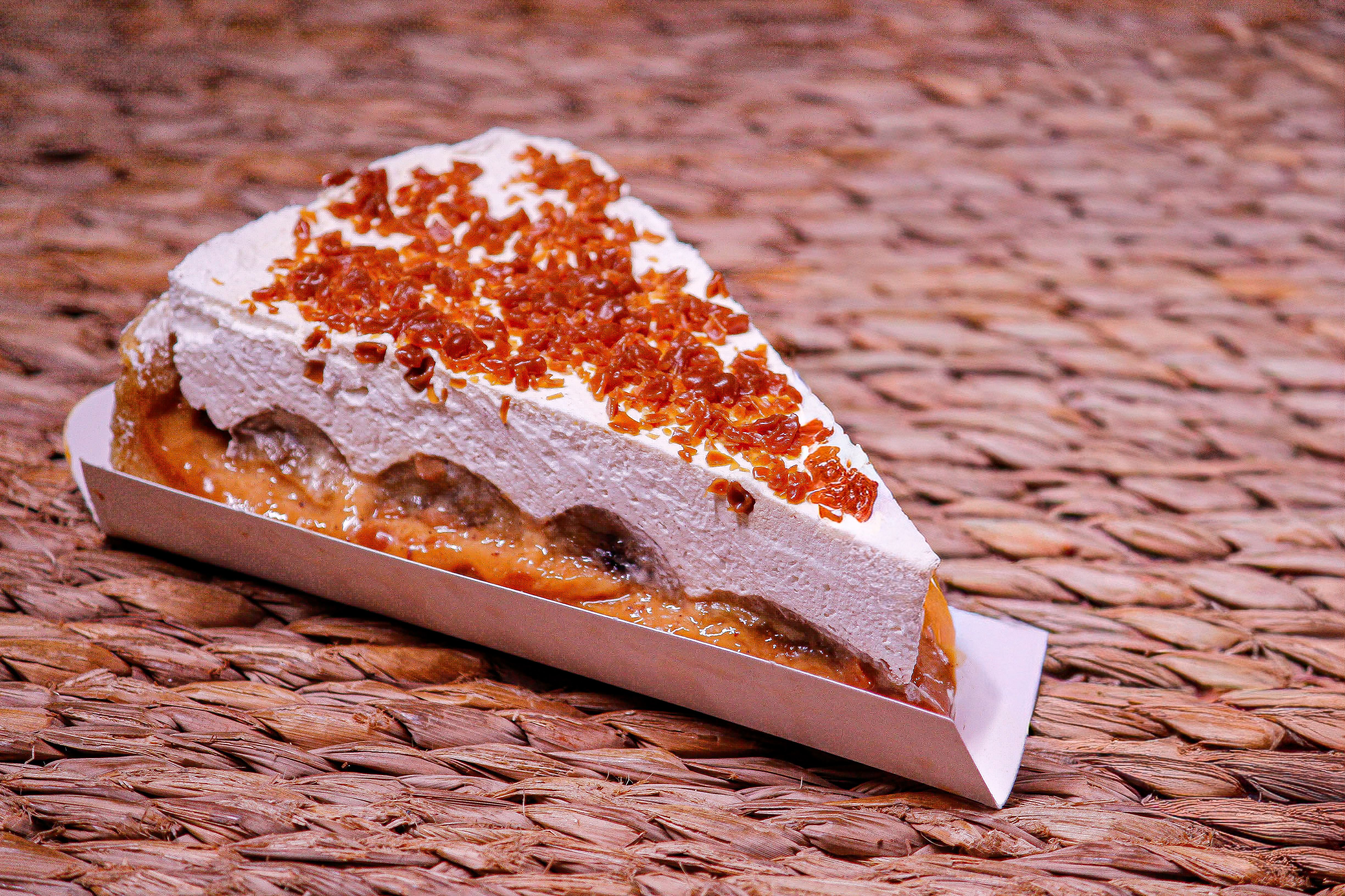 Fatia de torta de coco com calda de ameixa e cobertura de açúcar caramelizado, sobre uma superfície de fibra natural.