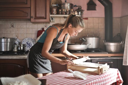 Free Side view of housewife wearing apron standing at table in cozy kitchen and preparing dough for baking while using rolling pin Stock Photo