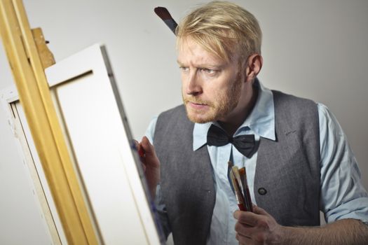 Focused male artist in elegant outfit creating artwork using canvas and easel while standing in workshop with paintbrushes