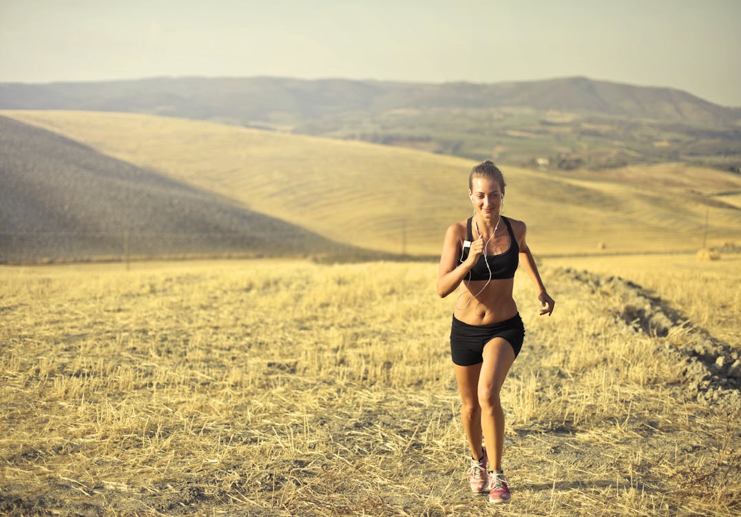 Free Powerful young female athlete in activewear running along hill on background of mountainous landscape and listening to music in earphones during cardio training Stock Photo