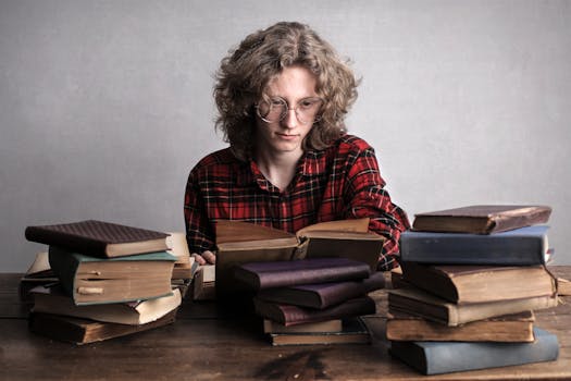 Smart male student with long curly hair wearing trendy checkered shirt sitting at table with piles of shabby books and reading literature in library