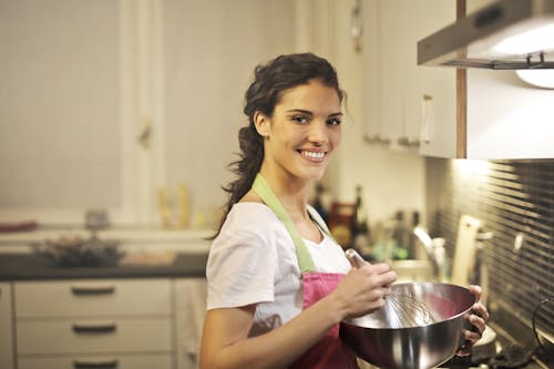 Side view of cheerful female in apron and casual t shirt standing in modern kitchen and mixing ingredients with whisk in stainless bowl while preparing dough