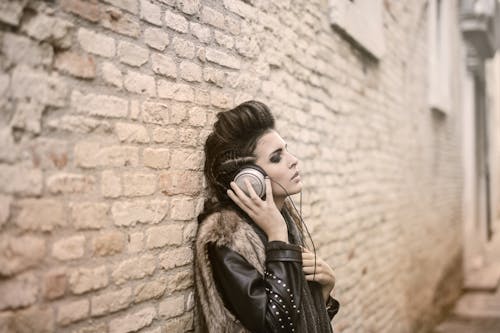 Free Side view of informal female in leather jacket and fur vest standing on street with closed eyes and enjoying songs in headphones while leaning on brick wall of building Stock Photo
