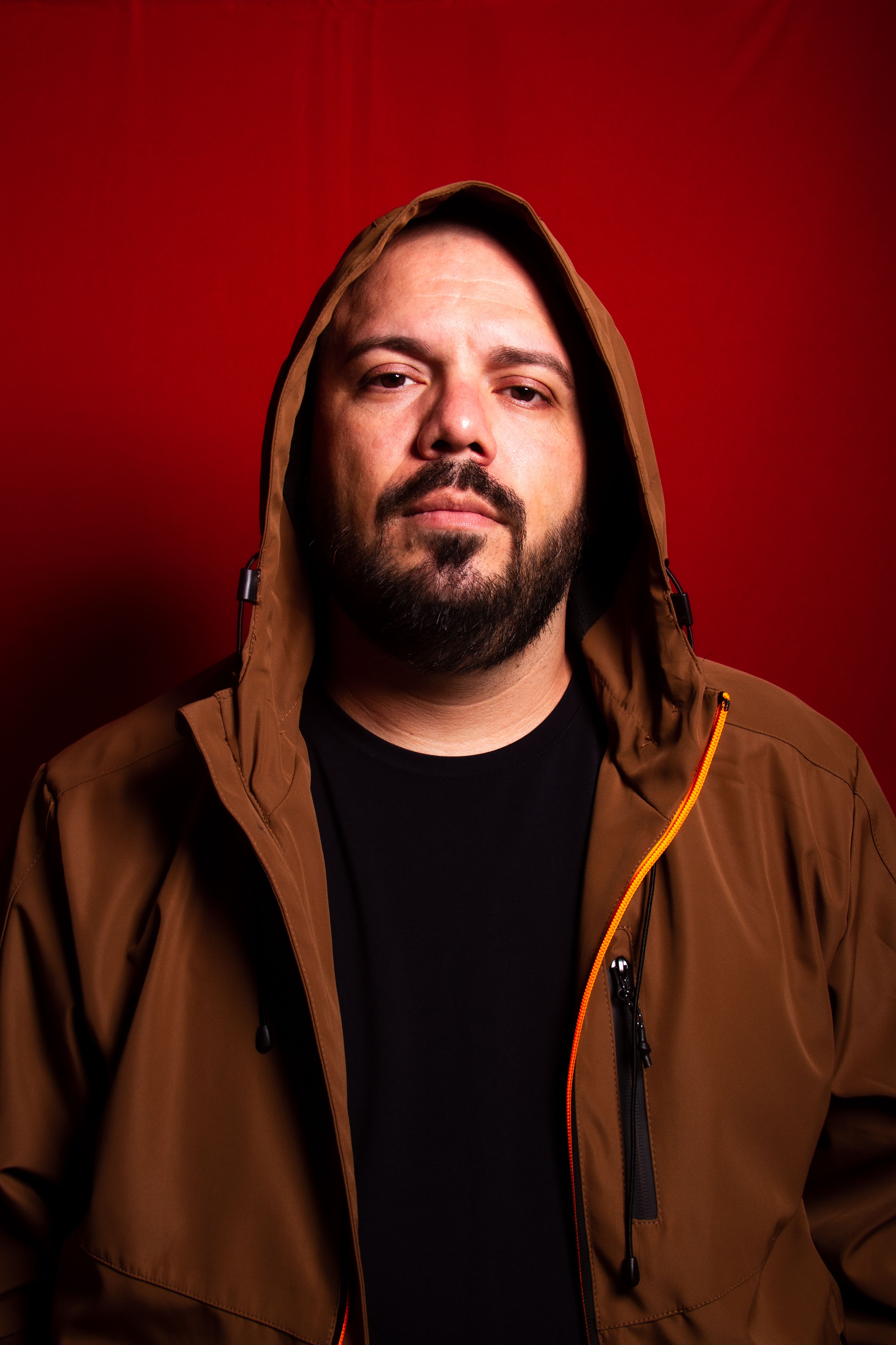 Expressive portrait of a bearded man in a hooded brown jacket with a red background.