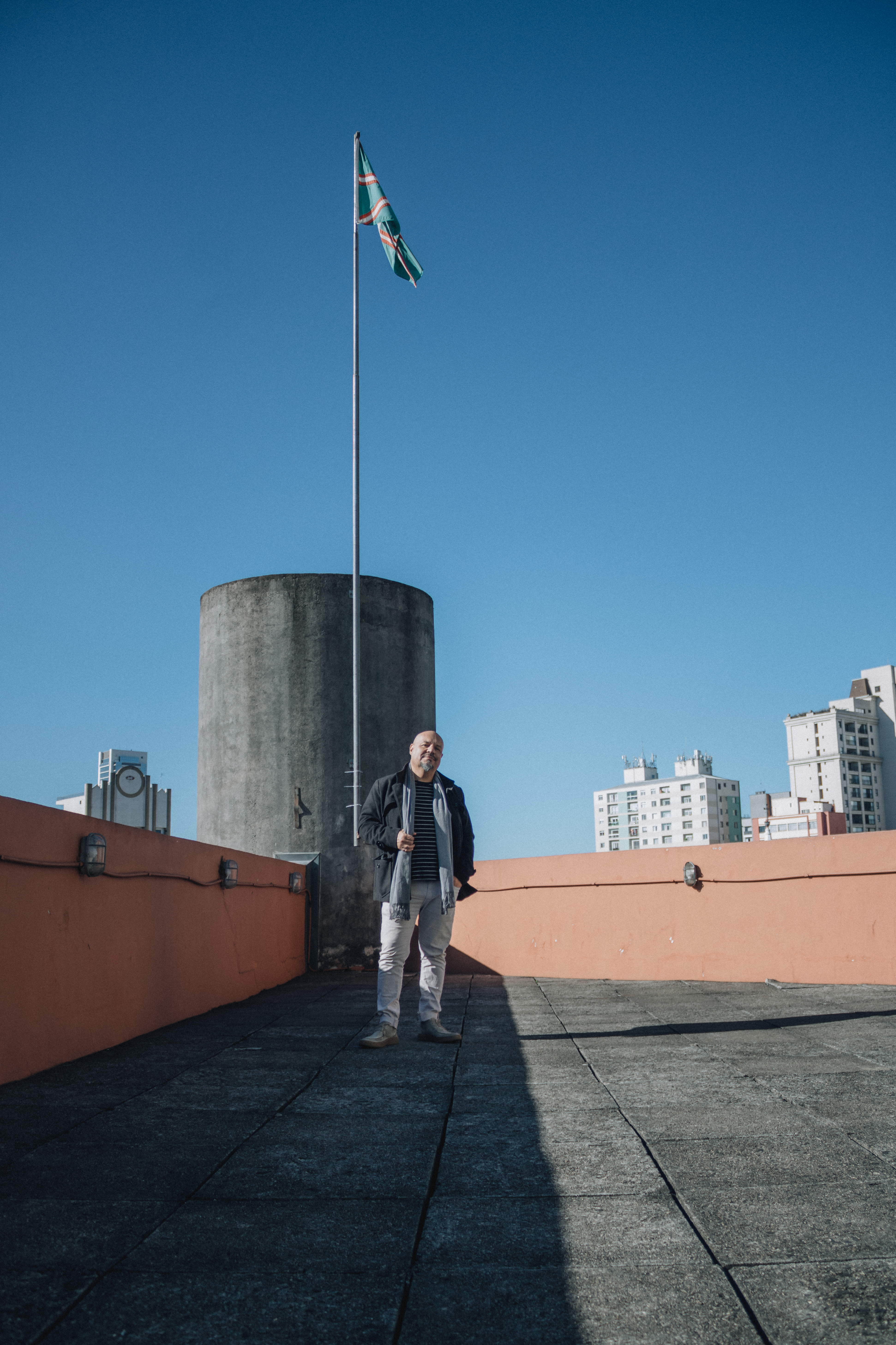 A man stands on a rooftop beside a flag, urban skyline in the background.