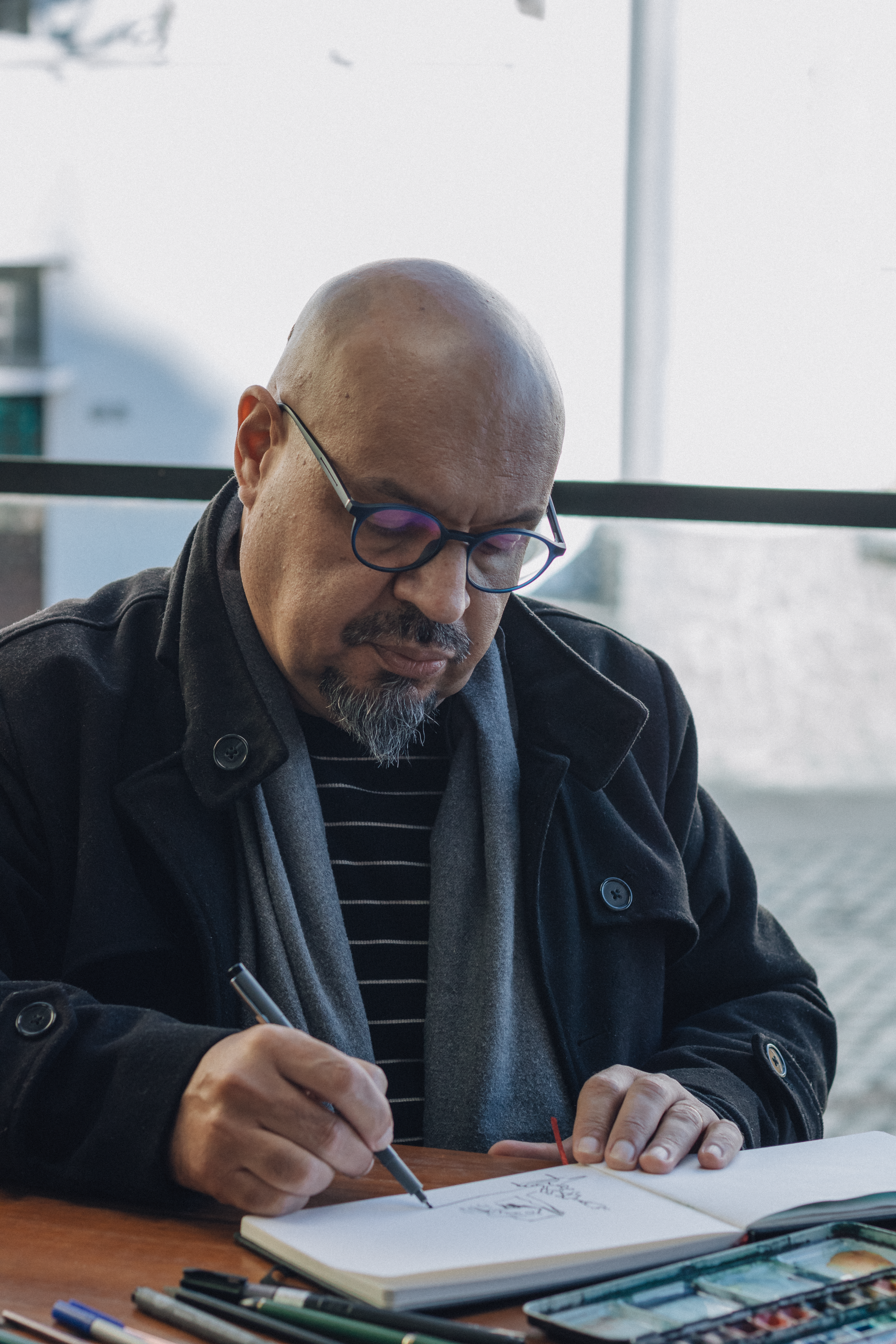 Middle-aged man sketching in a notebook by a window, wearing glasses and a coat.