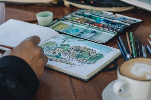 Free An artist sketching a landscape in a notebook with watercolors, coffee nearby. Stock Photo