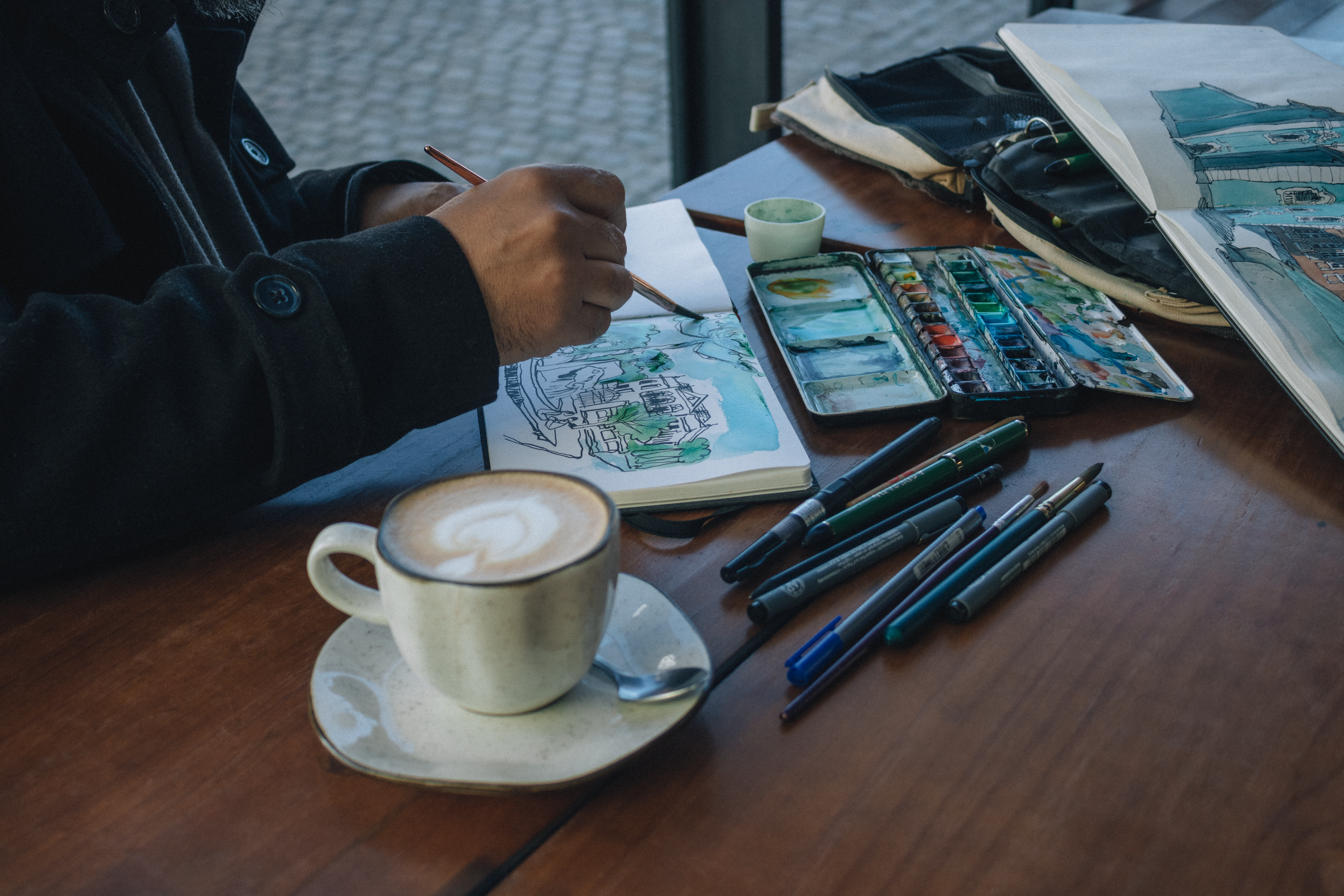 Artist drawing in a cafe with coffee, using watercolors and pens on a sketchbook.