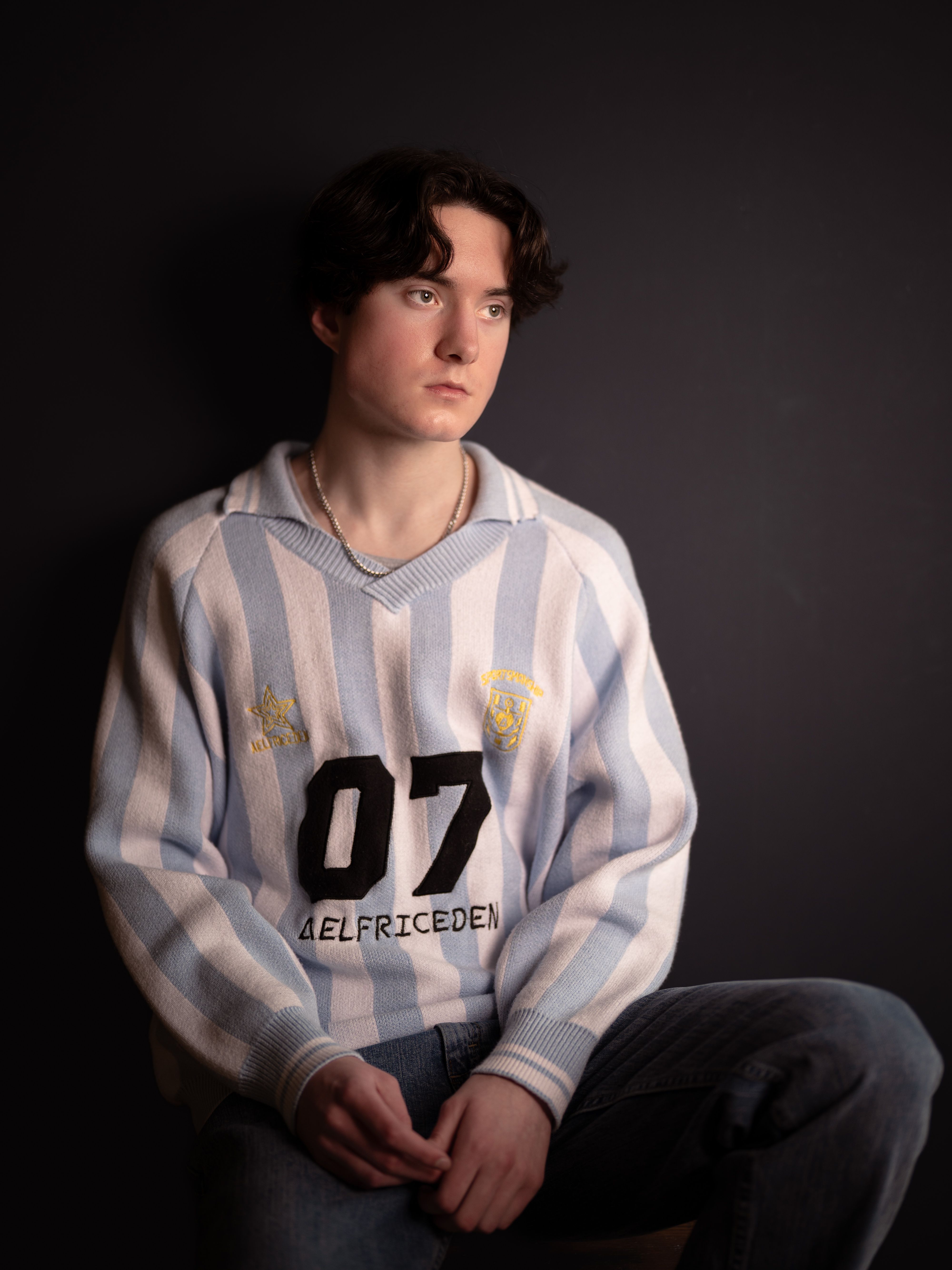 Contemplative young man in a striped sweater sits in a dimly lit room.