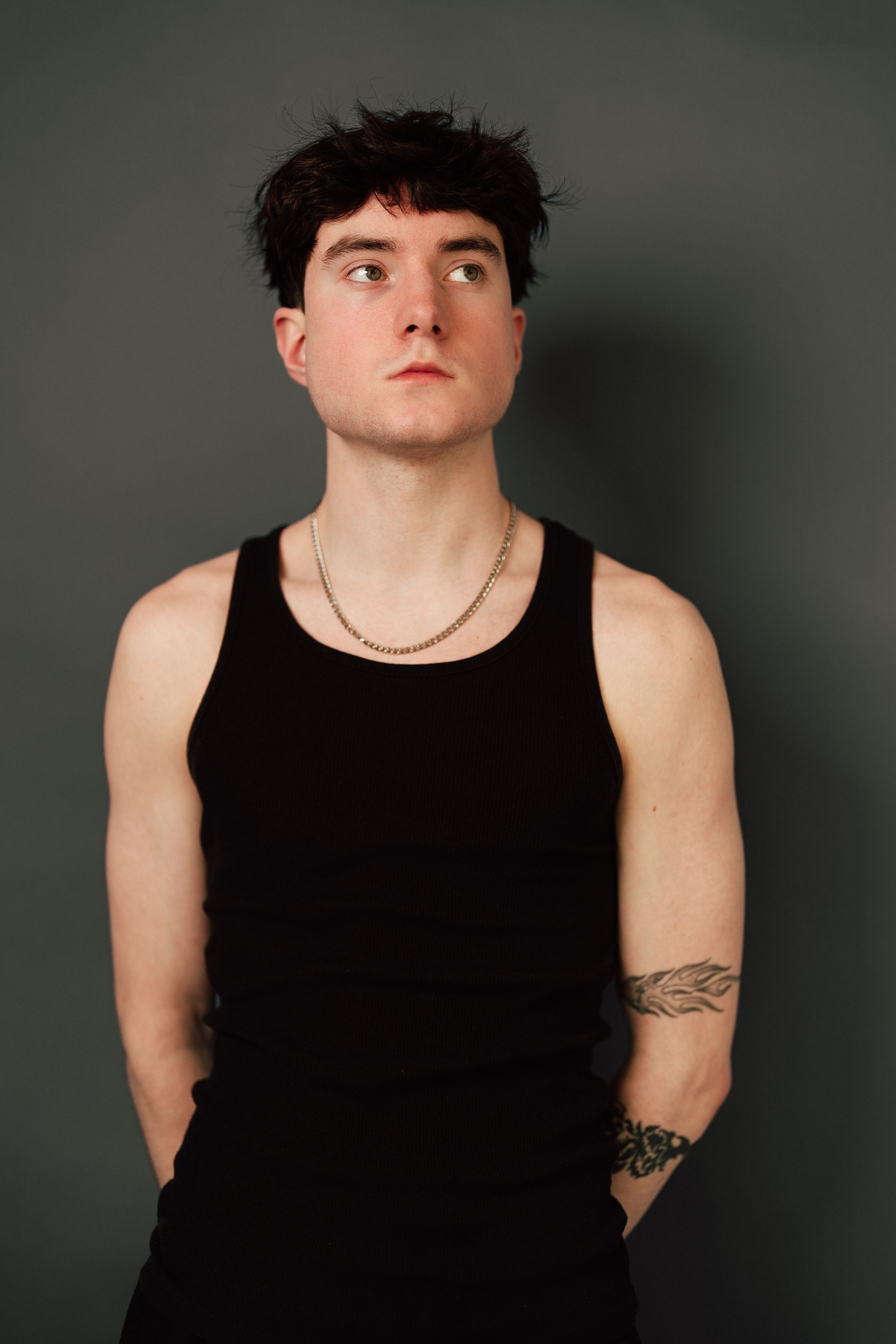 A thoughtful young adult man in a black tank top poses against a minimalist backdrop.