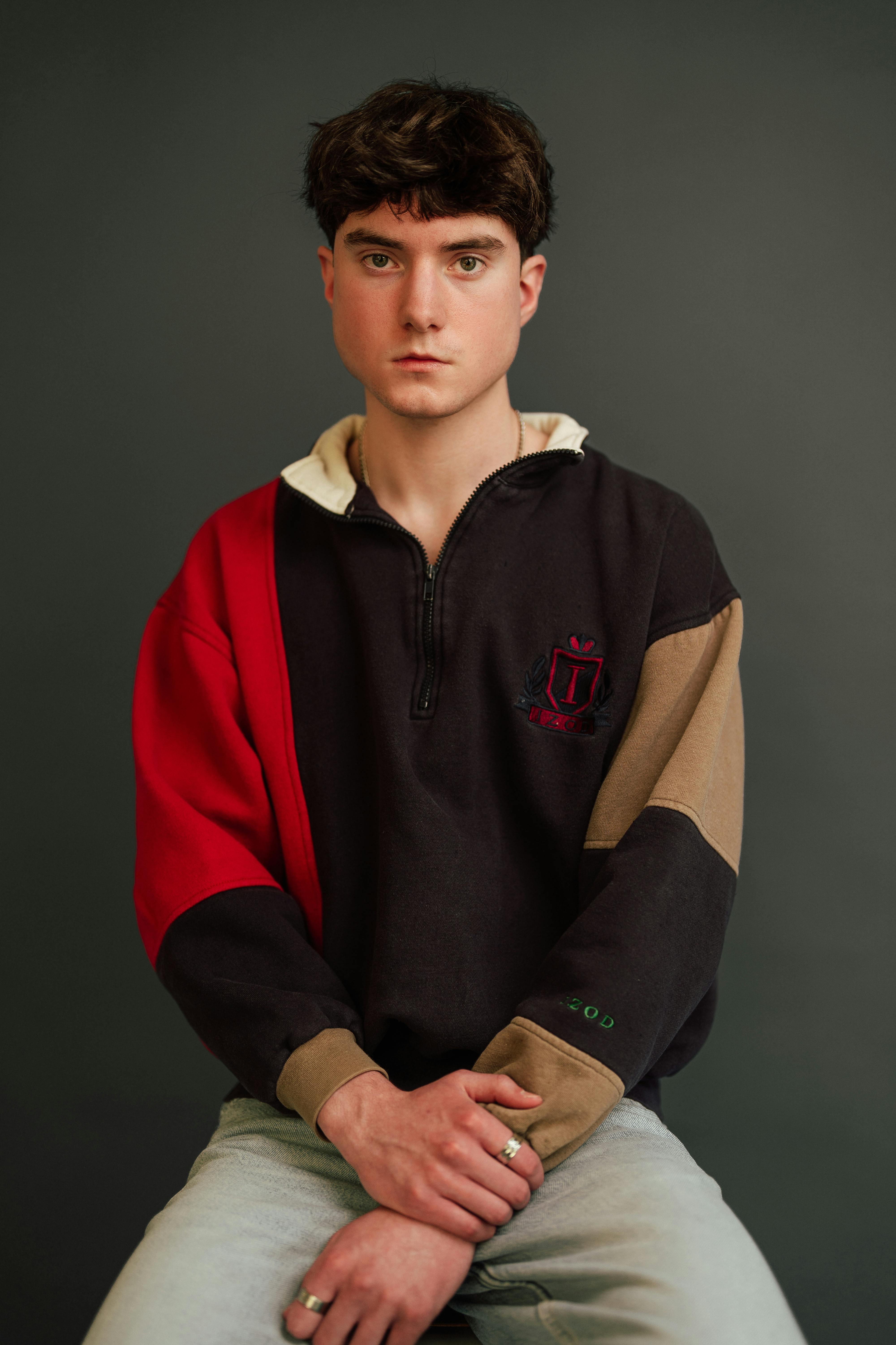 A young adult seated in a colorful sweater against a dark backdrop, looking directly at the camera.