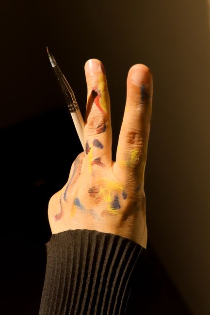 An artistic hand holding a paintbrush, fingers marked with vibrant paint