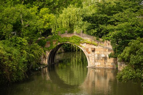 Free A scenic view of an ancient stone bridge surrounded by lush greenery, reflecting in a calm river. Stock Photo