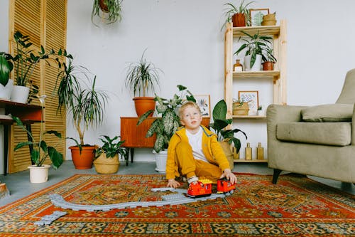 Playful child in casual wear sitting on carpet in cozy living room and playing with plastic railway and train while having fun during weekend