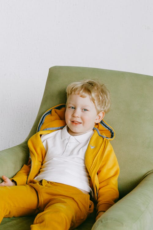 From above of cute smiling child in bright yellow tracksuit sitting in cozy armchair in living room  and looking at camera