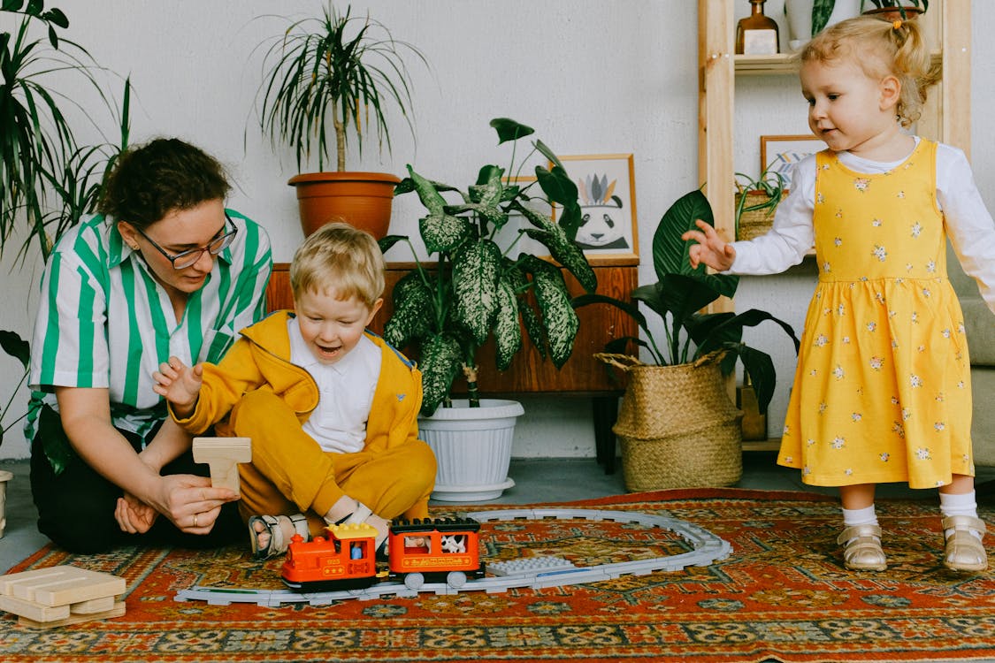 Free Mom and adorable little brother and sister in casual wear gathering in cozy living room during weekend and having fun together while playing with plastic railway Stock Photo