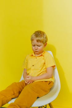 一个穿着黄色衣服的小男孩，坐在亮黄色的背景前，神情悲伤