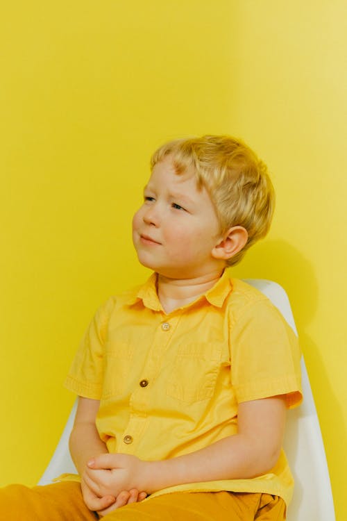 Boy in Yellow Button Up Shirt Sitting on White Chair Near Wall