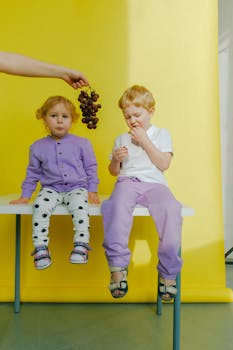 在黄色背景下，两个孩子坐在白色长凳上吃葡萄