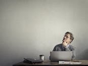 富有洞察力的商人坐在办公桌前，一边用笔记本电脑工作，一边喝着咖啡，这时他抬起了头