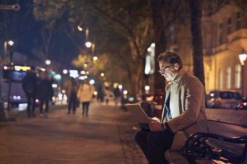 Man in Brown Coat Sitting on Bench Using Macbook 
