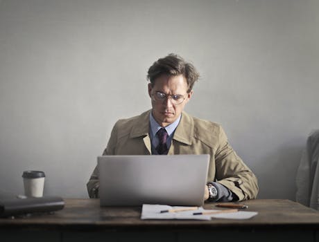 Man in glasses working intently on a laptop at a wooden desk indoors, showcasing concentration.