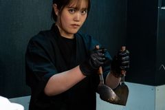 Female chef cooking fries on a stove in an indoor restaurant kitchen, wearing black attire and gloves.