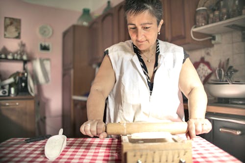 Aged female baker rolling gough with pin while cooking in cozy kitchen at home