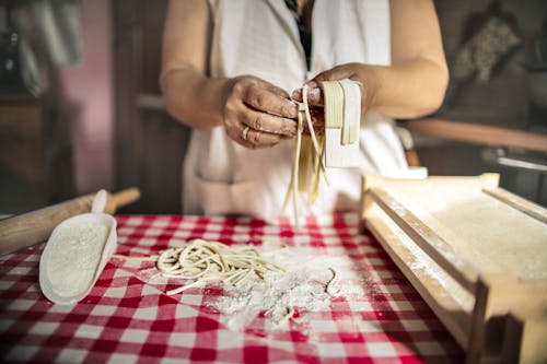 Unrecognizable cook in uniform standing at table doing noodles from dough in kitchen at home