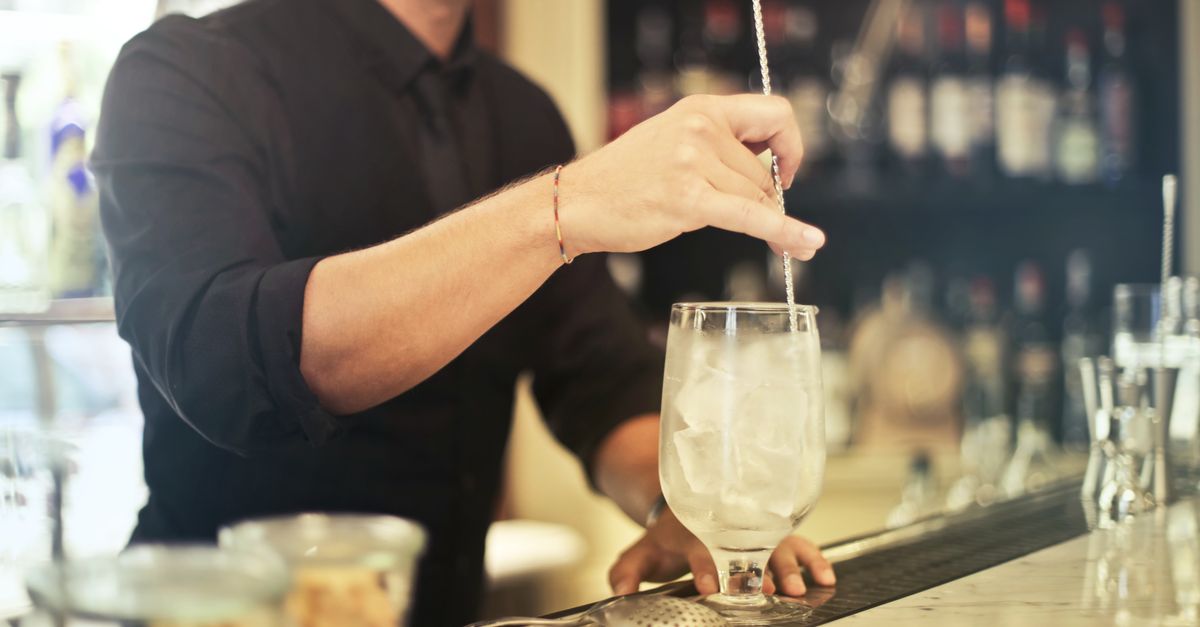 Faceless bearded male bartender in uniform making alcohol cocktail with ice in glass at bar