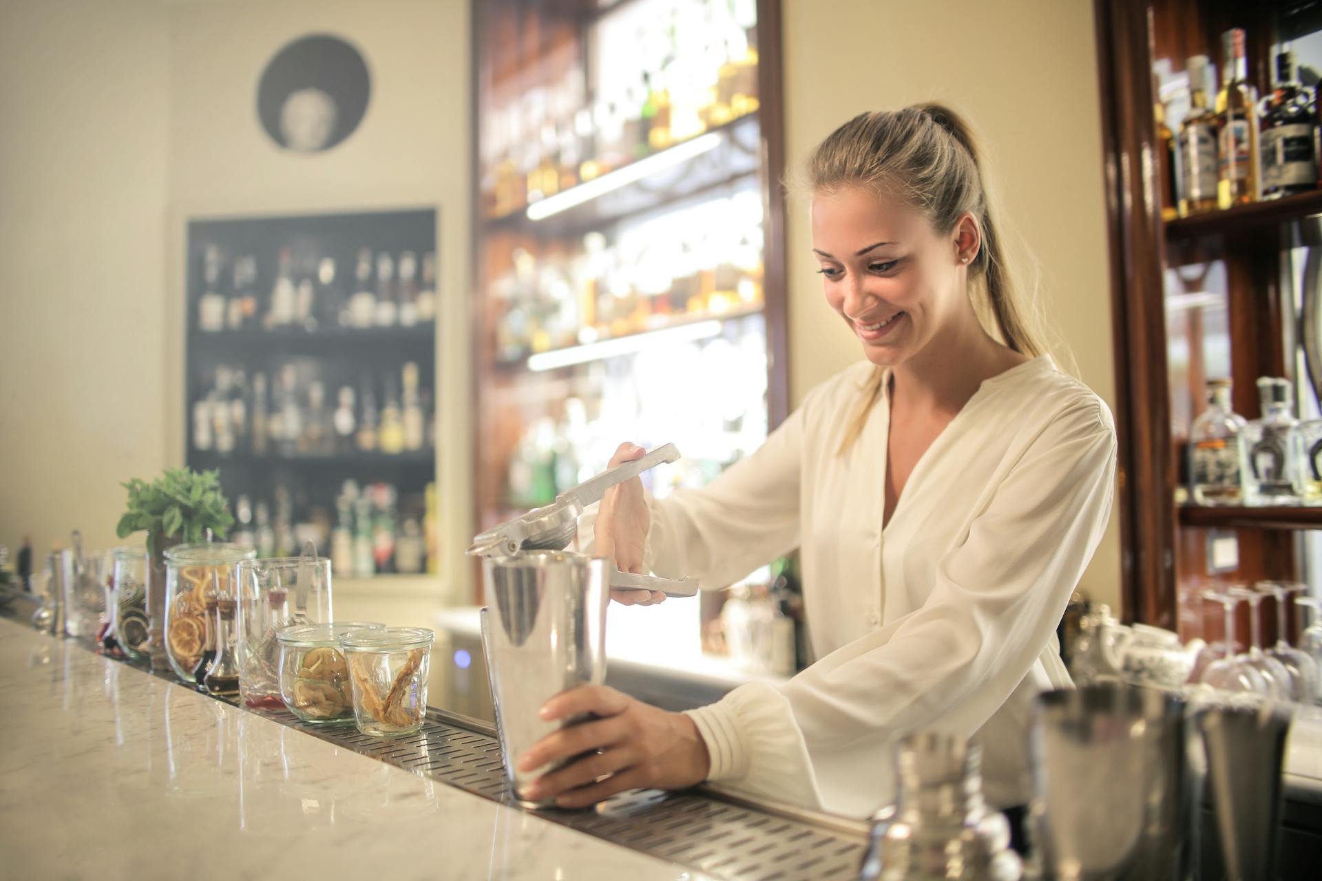 Smiling blonde in white blouse squeezing fresh juice into stainless shaker while preparing cocktail in bar