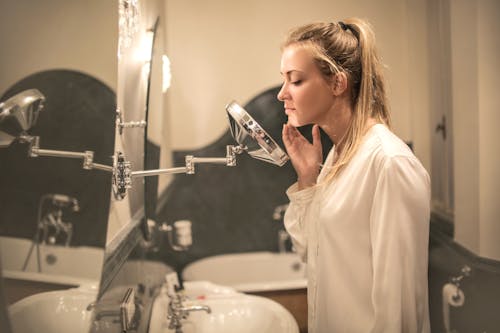 Free Side view of blond woman in white blouse standing in bathroom looking closely at face skin in small mirror Stock Photo
