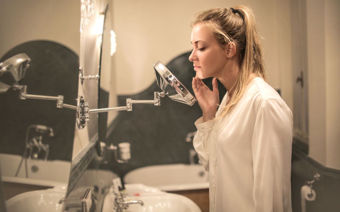 Woman in a white blouse looking closely at her face in a magnifying mirror in a bathroom