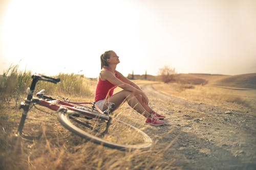 Full body of female in shorts and top sitting on roadside in rural field with bicycle near and enjoying fresh air with eyes closed