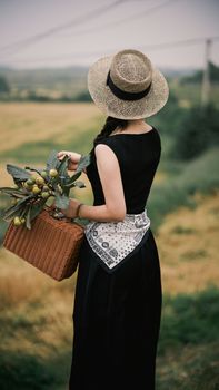 A woman in a straw hat holding a basket with fruit, facing a picturesque countryside view.