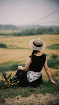 A serene scene of a woman in a black dress and straw hat sitting in a lush summer field.