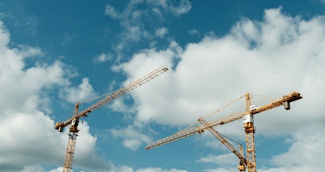 Two large tower cranes stand tall against a vibrant blue sky with fluffy clouds, symbolizing development.