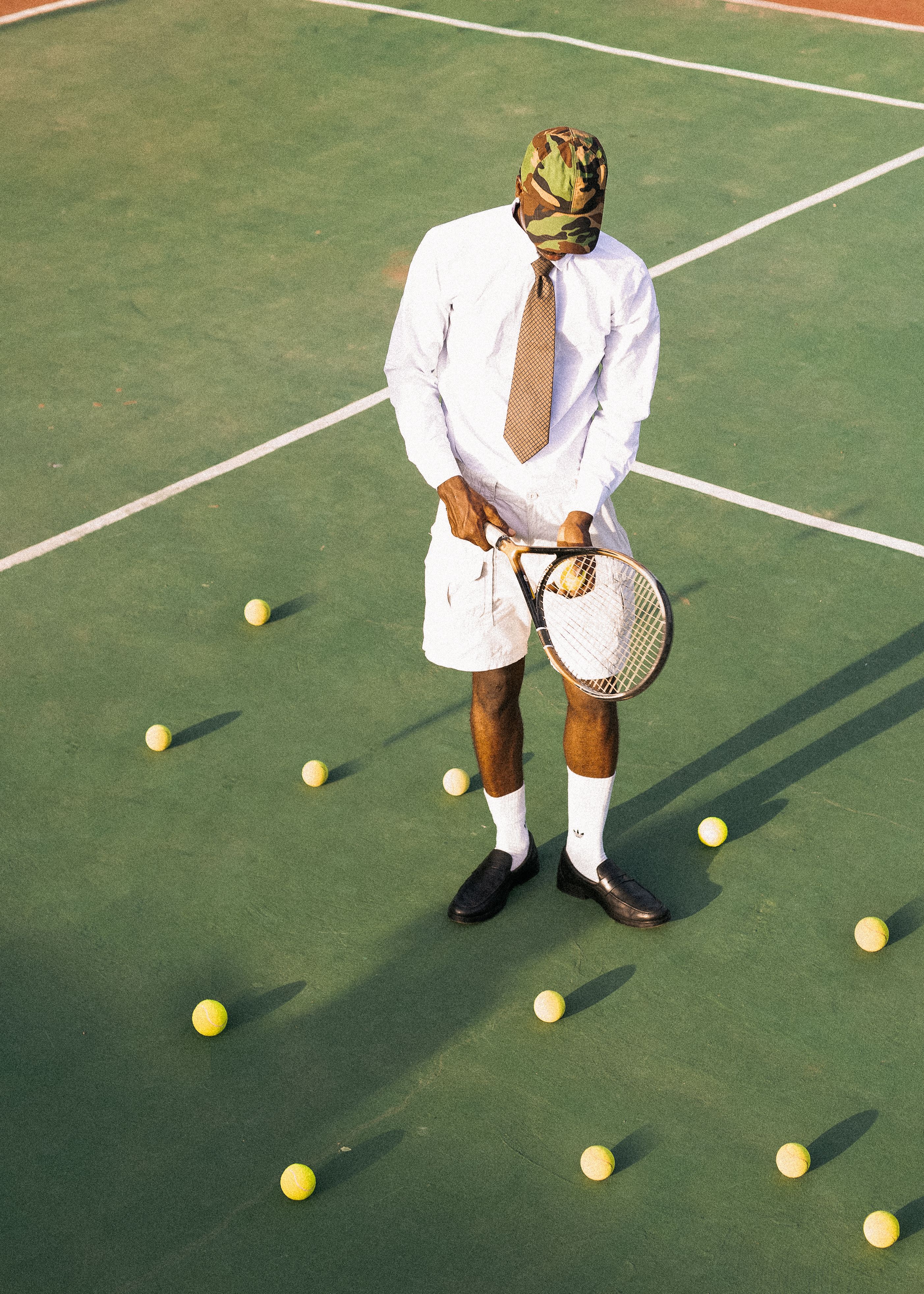 Man in formal attire with tennis racket on outdoor court surrounded by balls.