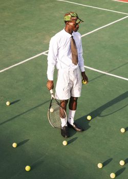 A fashionable man in formal attire holding a tennis racket on a tennis court surrounded by tennis balls.