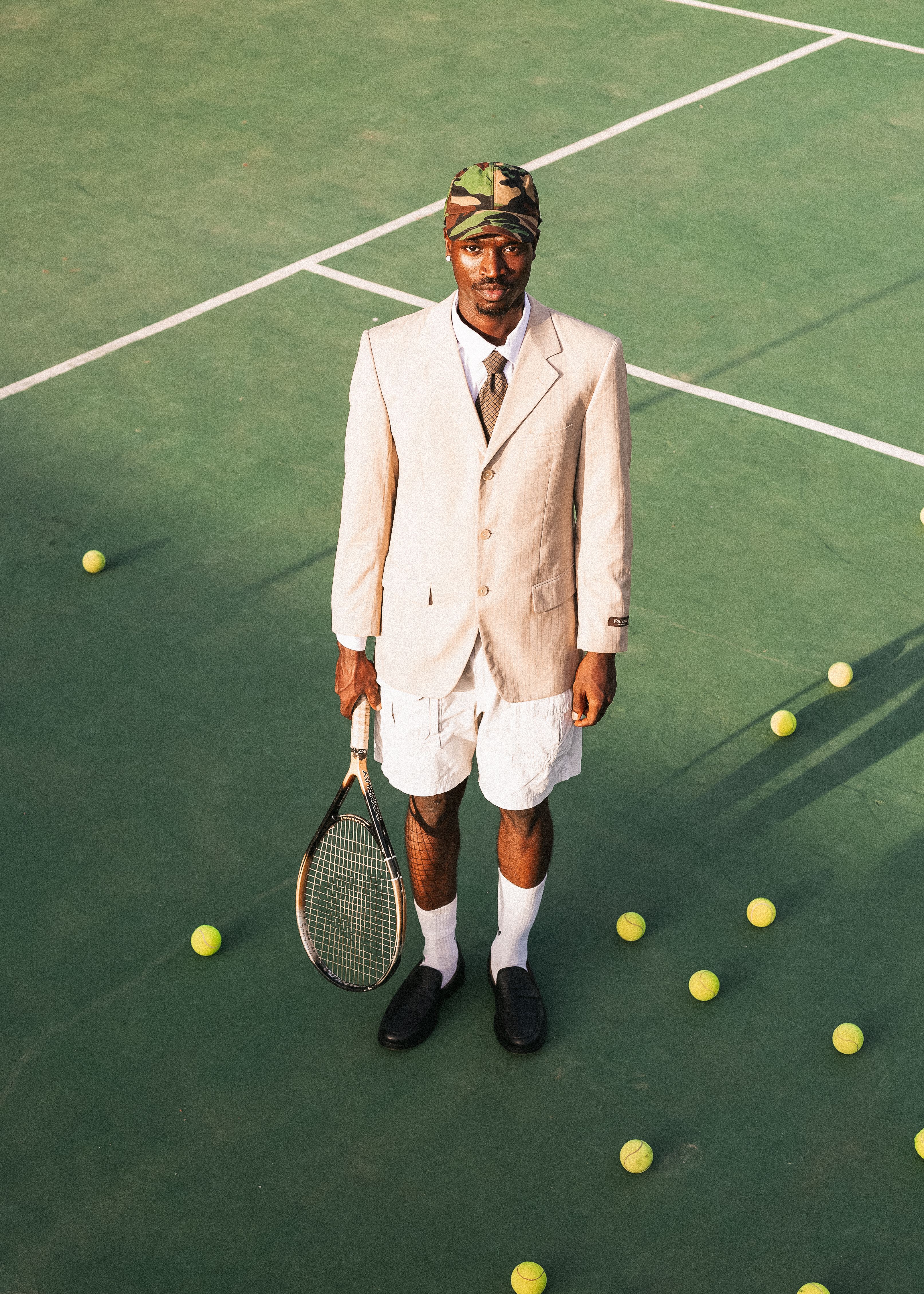 Man in blazer and shorts holding tennis racket on outdoor court surrounded by balls.